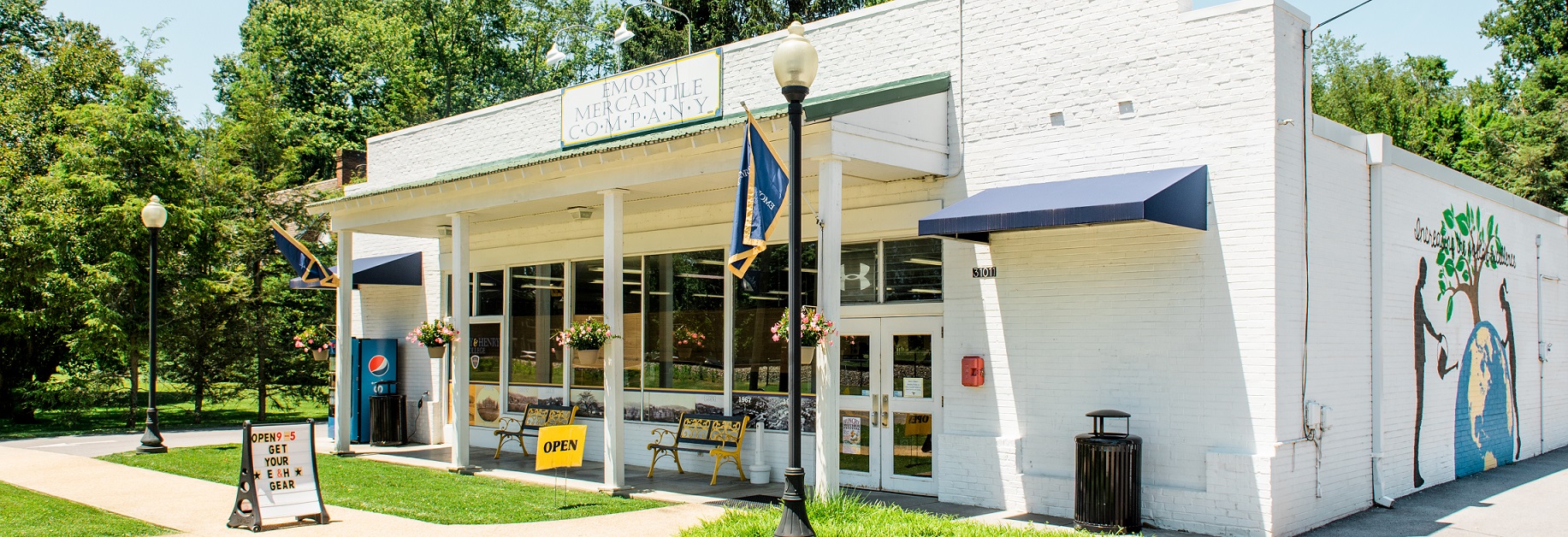 the exterior of the Mercantile at Oxford Ave, a white brick building with two benches and an Emory & Henry flag out front